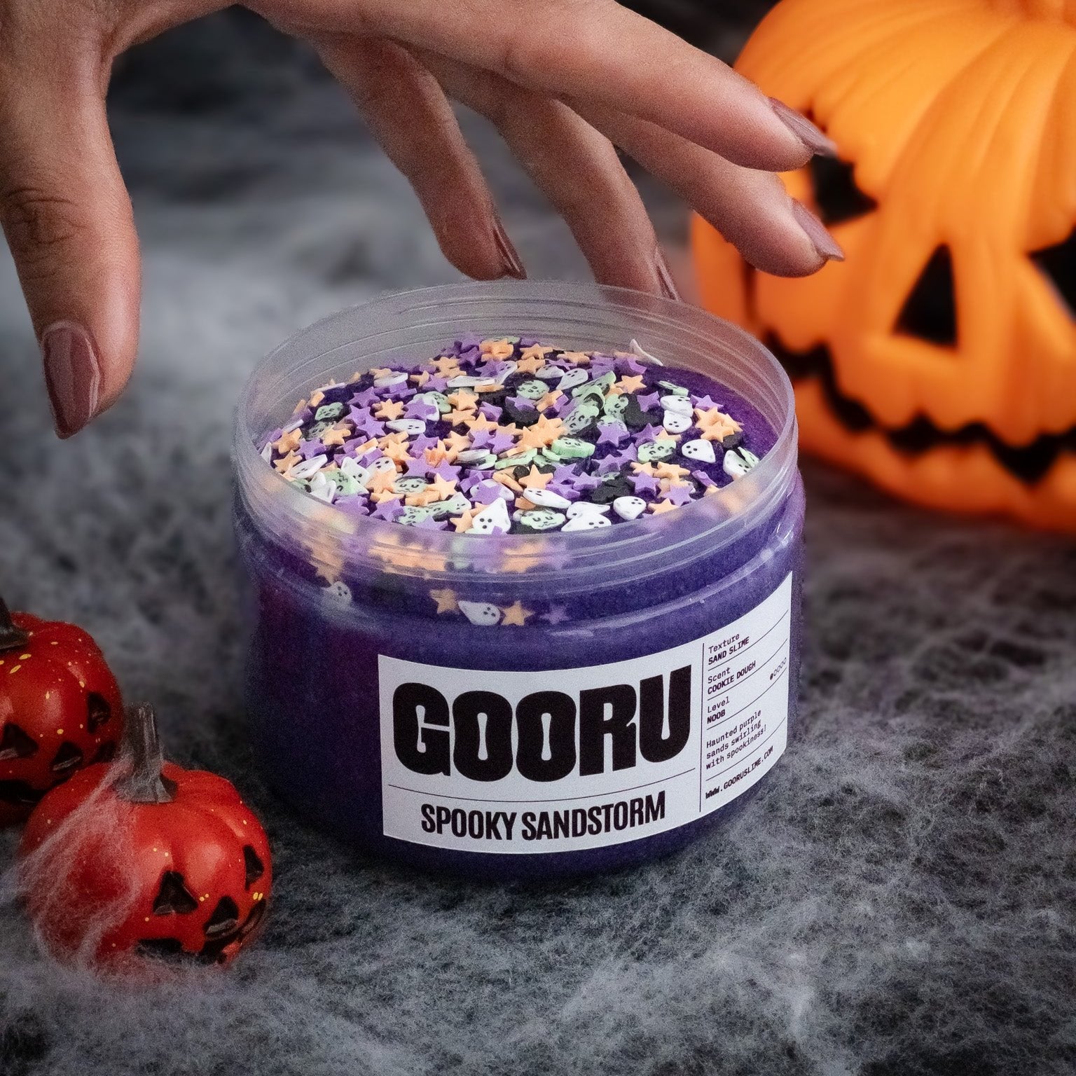 A tub of orange Jack-O-Floam slime with squishy foam beads, surrounded by spooky Halloween decorations like jack-o'-lantern ornaments and cobwebs. A hand is reaching to grab the slime tub, with wiggly googly eyes peeking through the foam, adding a playful and eerie vibe to the Halloween scene.