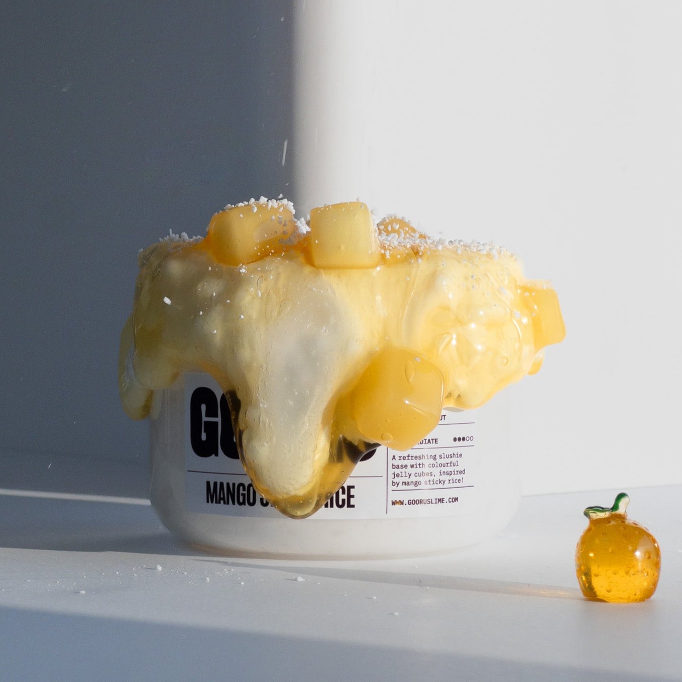 A jar of Mango Sticky Rice slime with white base and mango colored clear slime and a mango charm on the side with coconut sprinkles falling down on the slime