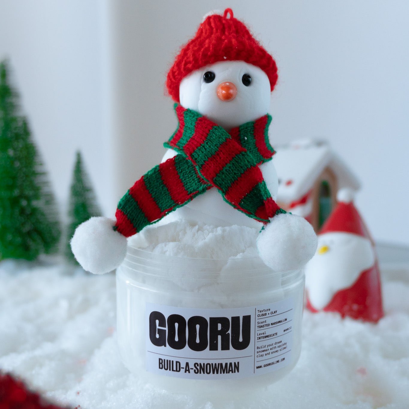  The Build-A-Snowman slime displayed in its festive setting, featuring the snowman with its red hat and striped scarf atop fluffy white slime. A cheerful Christmas scene with small trees and holiday decorations surrounds the jar.