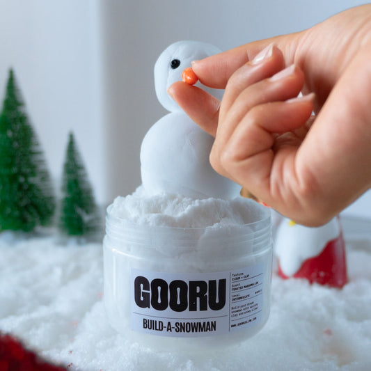 A creative moment of assembling the Build-A-Snowman slime set, with a hand carefully placing an orange carrot nose on a white clay snowman figure. The snow-like slime and holiday-themed decorations complete the scene.