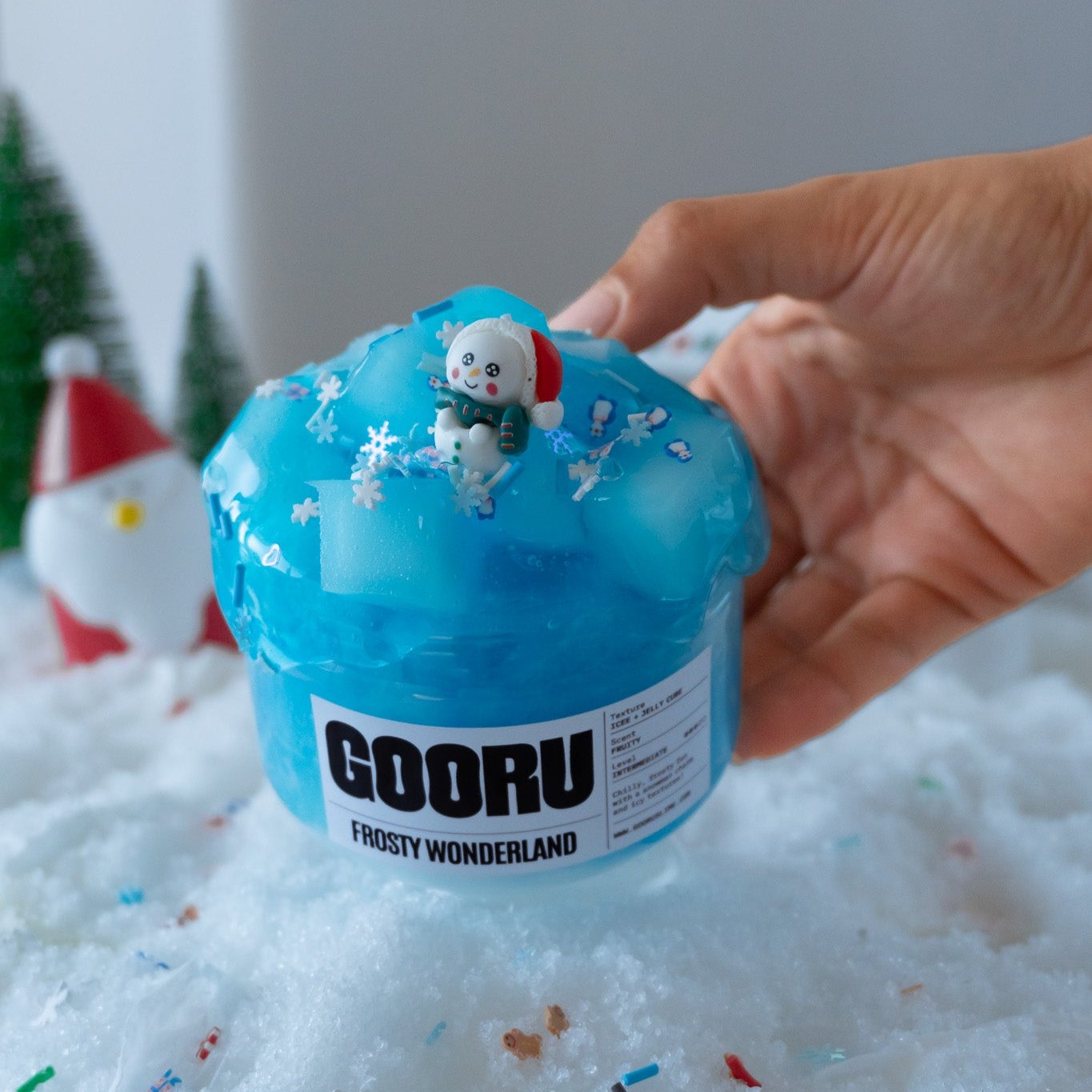 A close-up of Frosty Wonderland slime, showcasing its glossy blue texture topped with jelly cubes and snowflake charms. A hand holds the jar, highlighting the adorable polar bear figurine in a Santa hat.