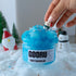 A playful interaction with Frosty Wonderland slime as a hand places the polar bear charm on the jelly cube-filled, bright blue slime. The snowy setup and festive decorations create a cheerful winter atmosphere.