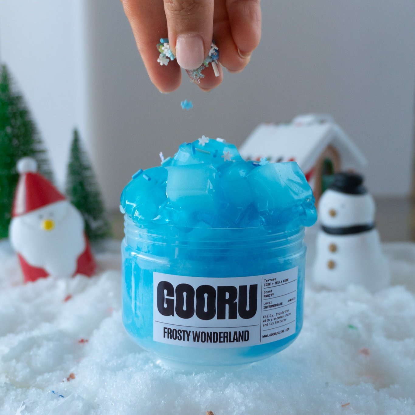 A jar of Frosty Wonderland slime featuring a vibrant blue color with jelly cube textures, surrounded by a snowy setup. A hand is sprinkling snowflake-shaped charms onto the slime, adding a magical winter vibe.
