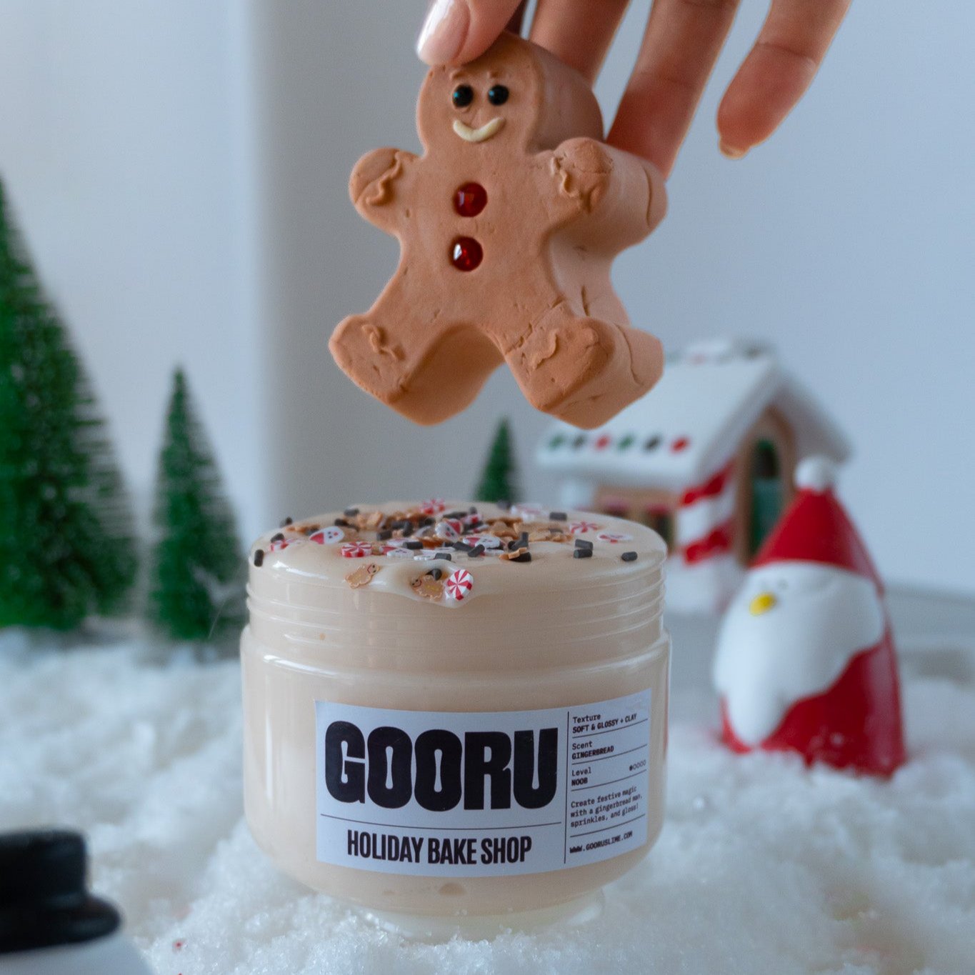 A festive jar of Holiday Bake Shop slime, featuring a soft and glossy texture with gingerbread-themed sprinkles on top. A hand is holding a cute gingerbread man clay charm above the jar, ready for play.