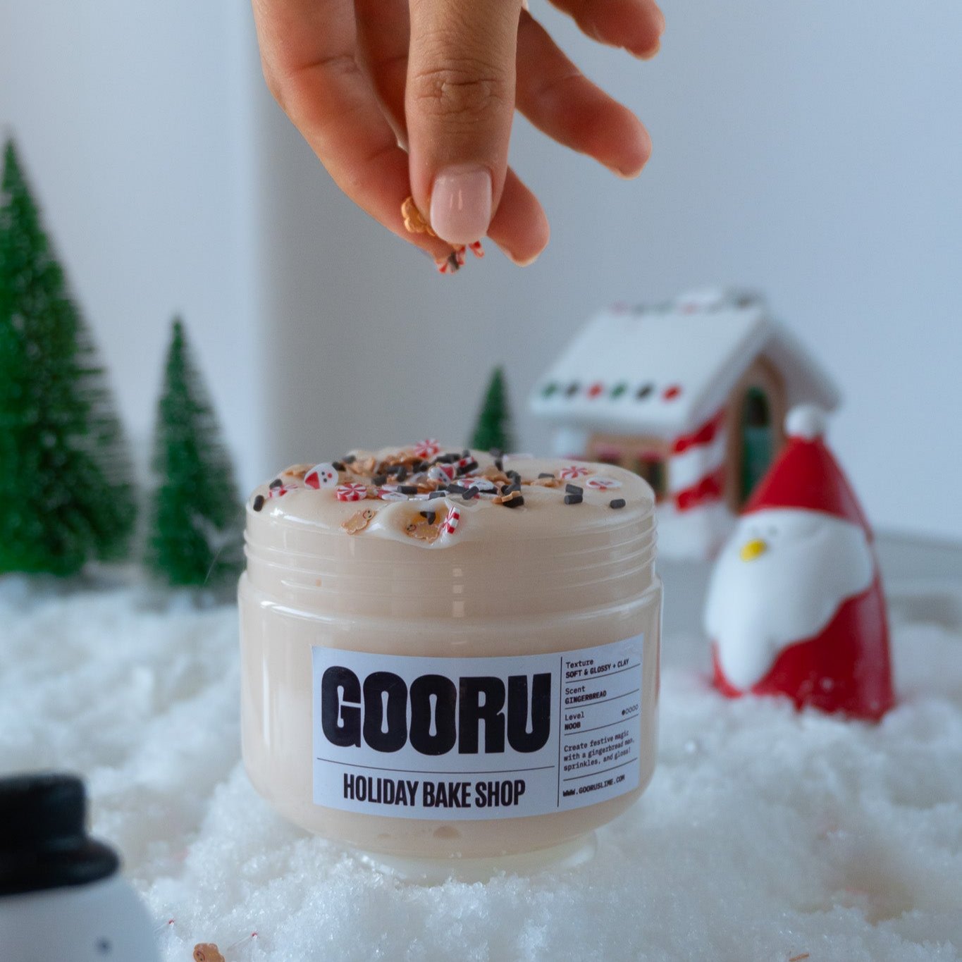 A hand sprinkling tiny holiday-themed decorations onto the creamy, gingerbread-scented slime in the Holiday Bake Shop jar. The festive background includes snow, Santa figurines, and Christmas trees.