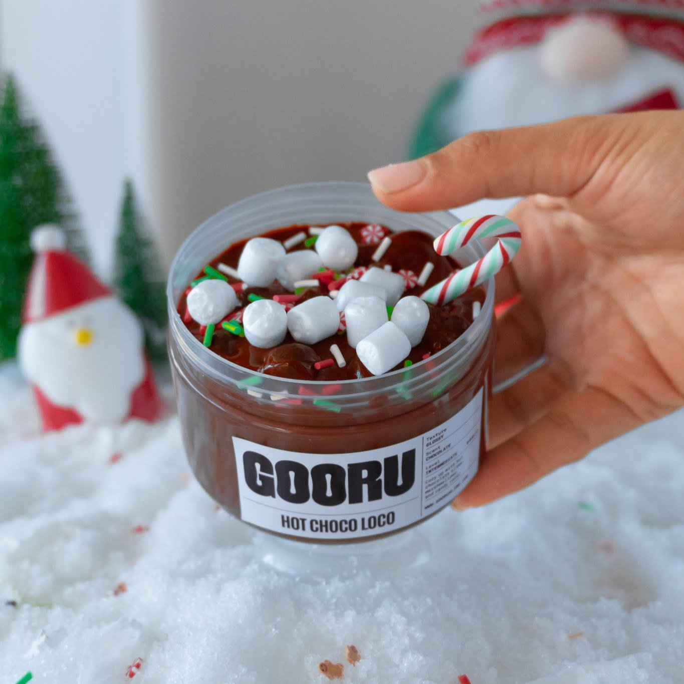 A jar of Hot Choco Loco slime with a glossy chocolate base, topped with mini marshmallows and a candy cane charm. A hand holds the jar, showcasing the cozy holiday-themed slime in a festive snowy setup.