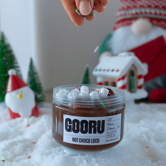  A hand sprinkling colorful holiday-themed sprinkles onto the glossy chocolate slime in the Hot Choco Loco jar. The scene is surrounded by Christmas trees and festive decorations, adding to the holiday vibe.