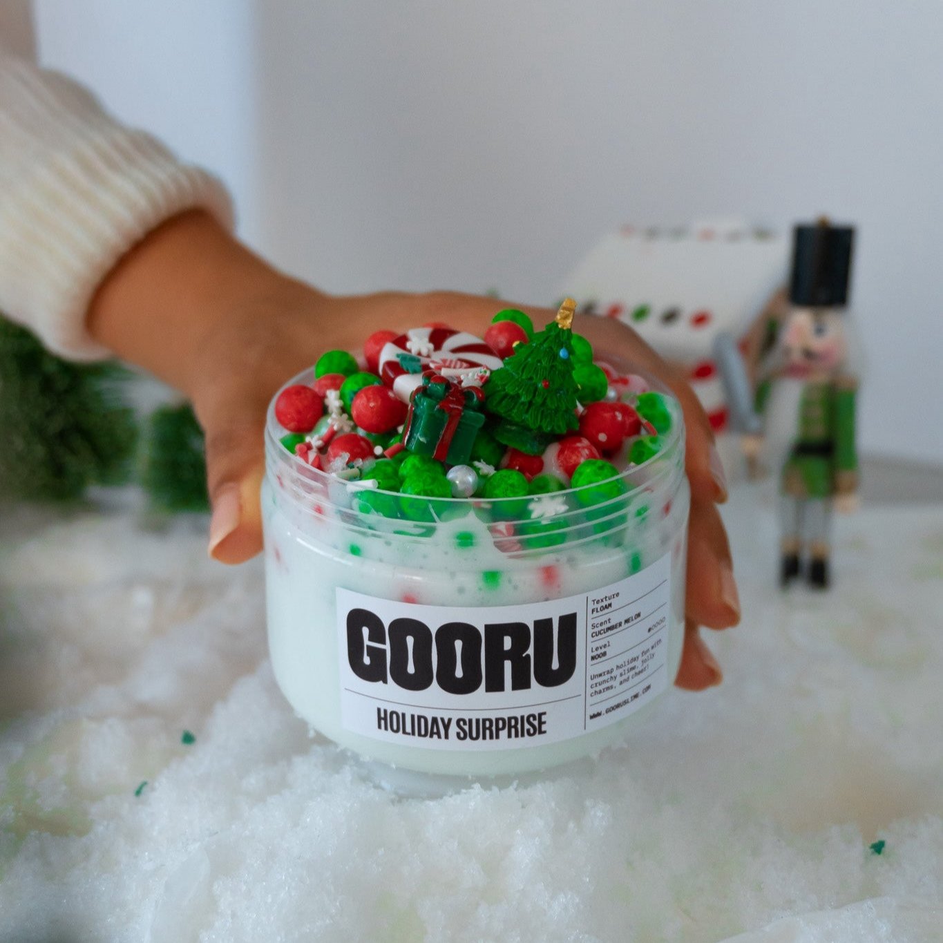 A hand holding the Holiday Surprise slime jar decorated with colorful foam beads, candy canes, and festive charms, including a Christmas tree. The background features a winter scene with a nutcracker and a gingerbread house.