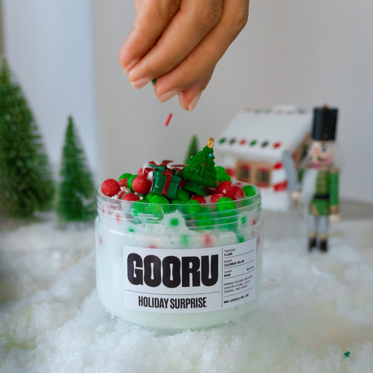 A jar of Holiday Surprise slime with a creamy white base and colorful foam beads, topped with festive charms. A hand is sprinkling holiday-themed decorations, adding to the cheerful Christmas aesthetic.