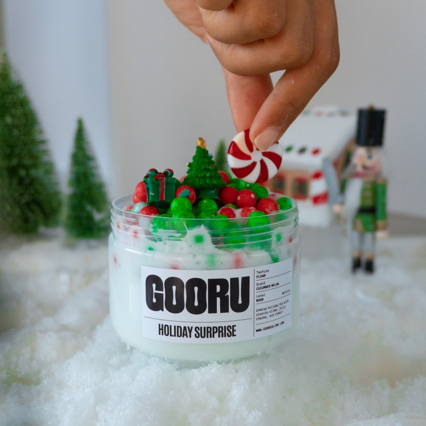 A playful interaction with Holiday Surprise slime as a hand places a red-and-white peppermint charm among the festive decorations. The jar is surrounded by a snowy holiday setup.