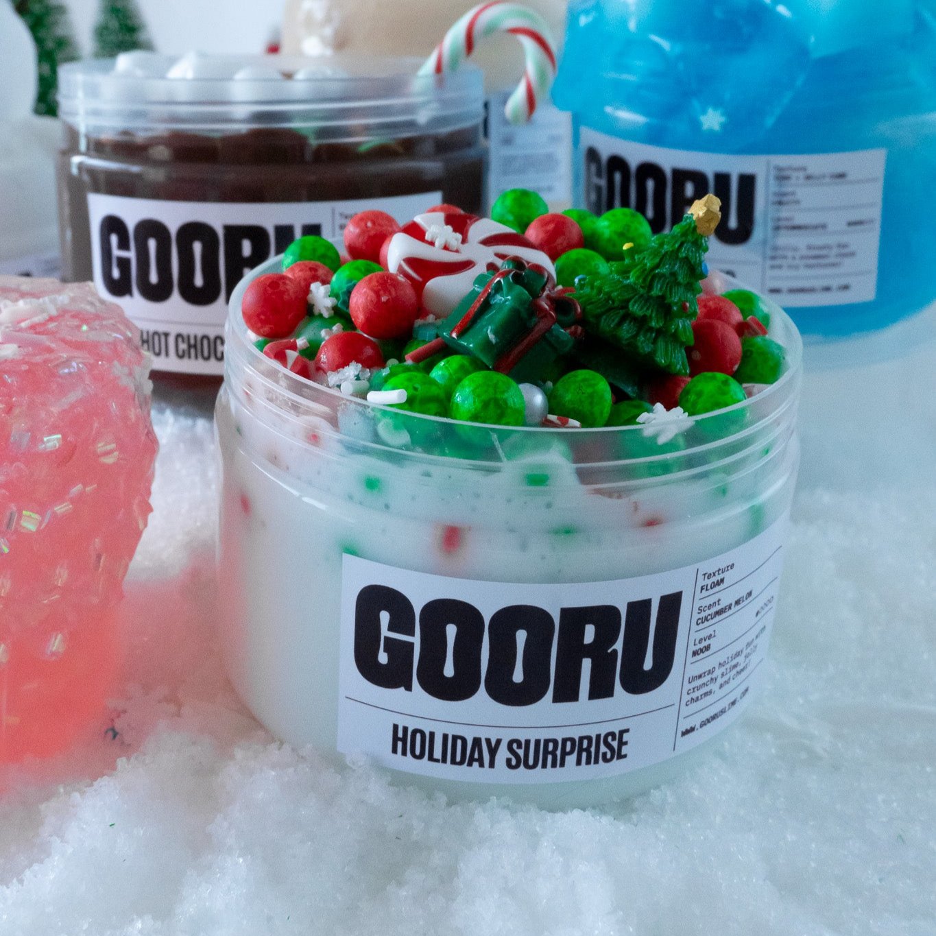 A group shot showcasing Holiday Surprise alongside other Gooru slime jars, including Hot Choco Loco and Frosty Wonderland. The Holiday Surprise jar stands out with its vibrant Christmas-themed decorations and cucumber melon scent.