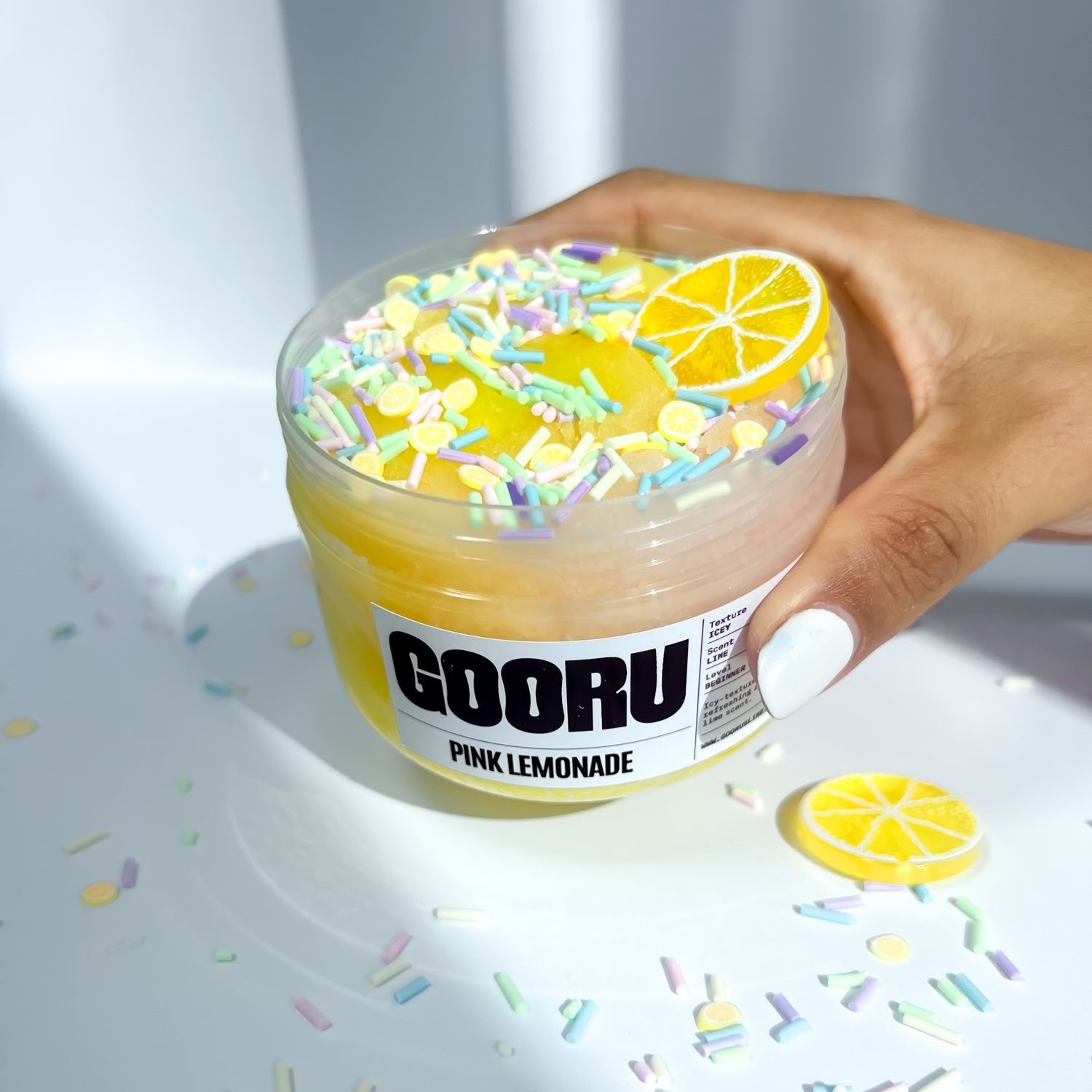 A product shot of  a hand holding Pink Lemonade slime, with lemon charm and colorful sprinkles, designed and manufactured  by Gooru Slime, Dubai, UAE