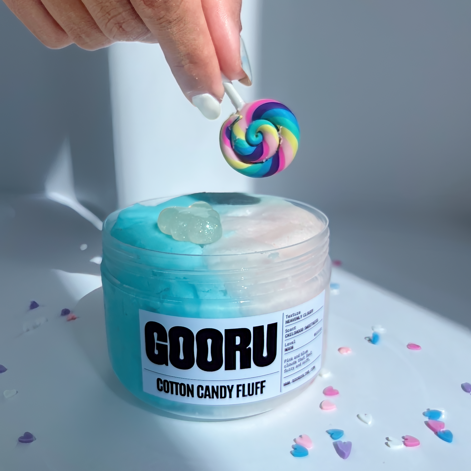 A product shot of Cotton Candy Fluff slime with hand placing a lollipop charm, designed and manufactured  by Gooru Slime, Dubai, UAE