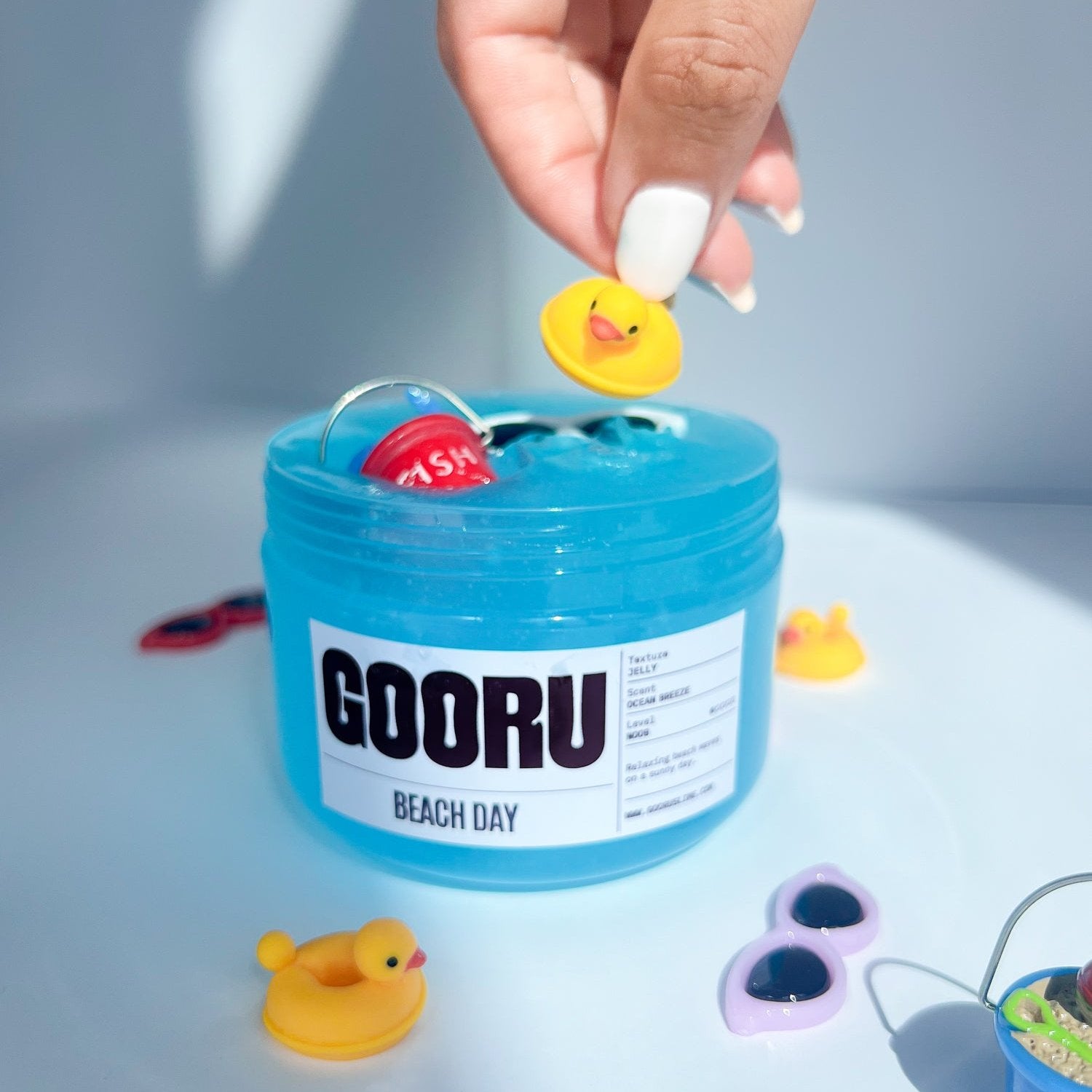 Product shot of hand holding a duck charm placing it into Beach day Slime, with beach bucket and sunglasses charms on top, designed and manufactured  by Gooru Slime, Dubai, UAE