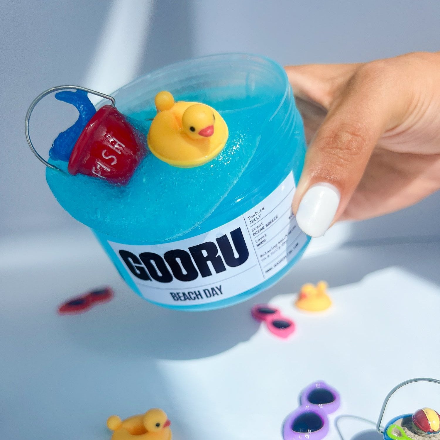 Product shot of hand holding Beach day Slime, with beach bucket and sunglasses charms on top, designed and manufactured  by Gooru Slime, Dubai, UAE