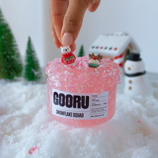 A hand placing a polar bear charm onto the pink, glittery Snowflake Squad slime. The snowy background and festive trees enhance the holiday-themed aesthetic.